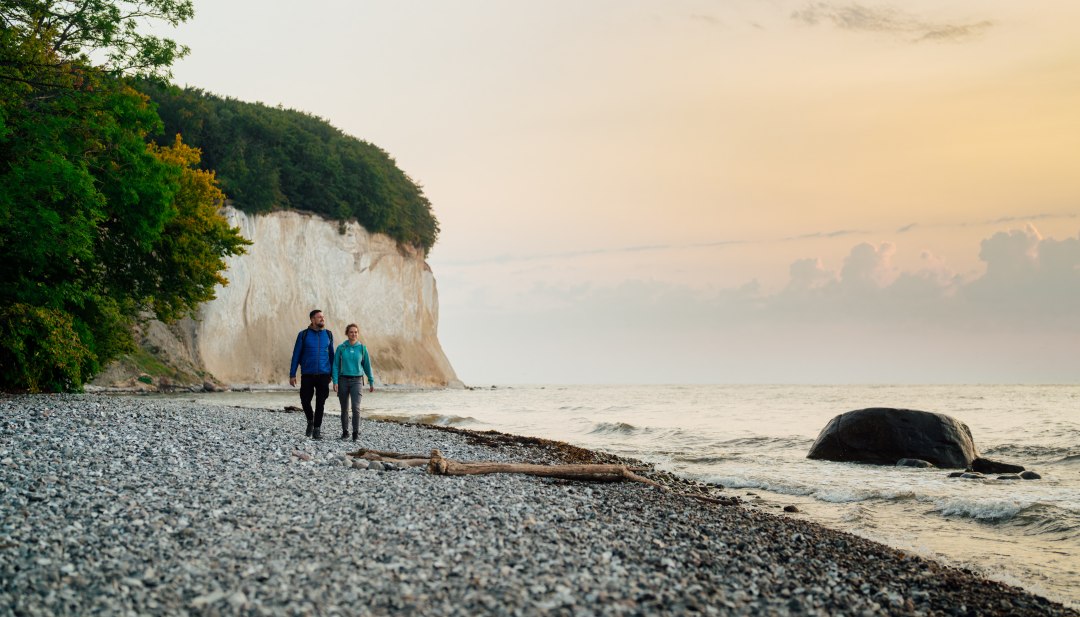 Een koppel wandelt langs de voet van de beroemde krijtrotsen op het eiland Rügen, terwijl de zonsondergang de hemel in zachte kleuren oplicht.