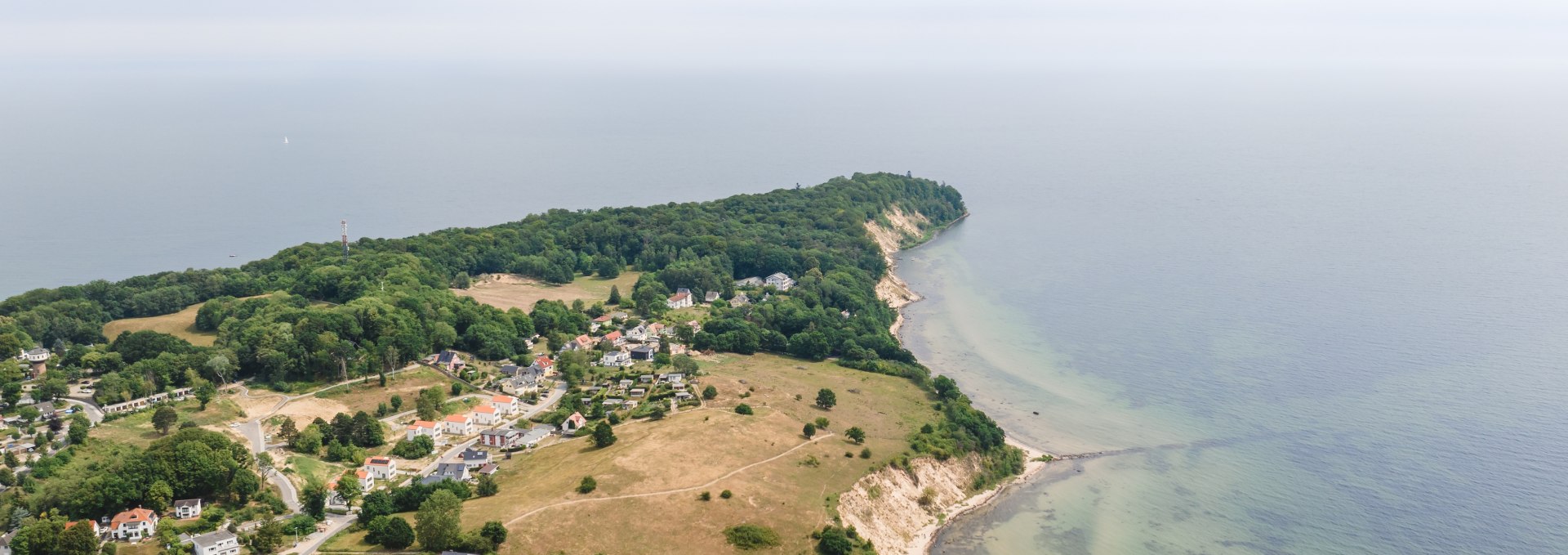 Widok z lotu ptaka na nadbałtycki kurort Göhren na wyspie Rugia z rozległymi łąkami, stromym wybrzeżem i Morzem Bałtyckim w tle pod lekko zachmurzonym niebem.
